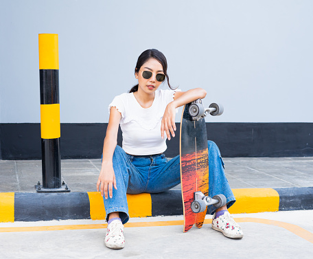 Asian cute girl or woman sit on street, place skateboard in vertical, and act smart in front of cool wall with copy space in daylight time, summer holiday.