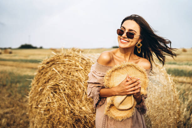 Smiling woman in sunglasses with bare shoulders on a background of wheat field and bales of hay stock photo