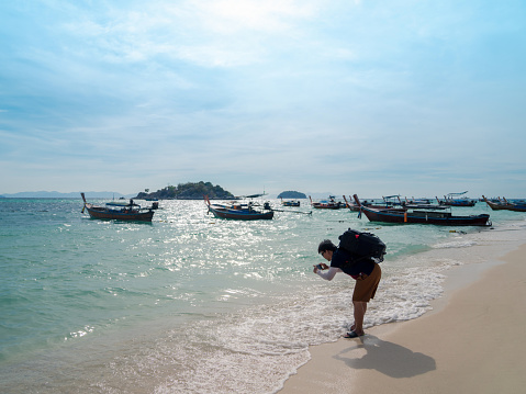 Asian backpacker tourist man taking photograph of sea wave with smartphone at sunny beach with sunlight reflection on water surface on Lipe island, Thailand, man relax on vacation in summer