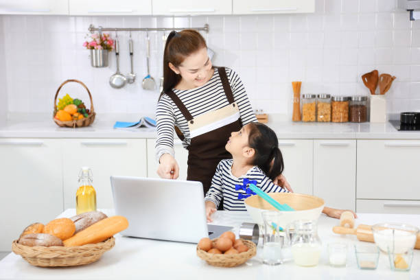 feliz madre asiática enseñando a su hija a hornear pan en la cocina moderna blanca mirando la receta en línea desde la computadora - bun bread 7 grain bread dough fotografías e imágenes de stock