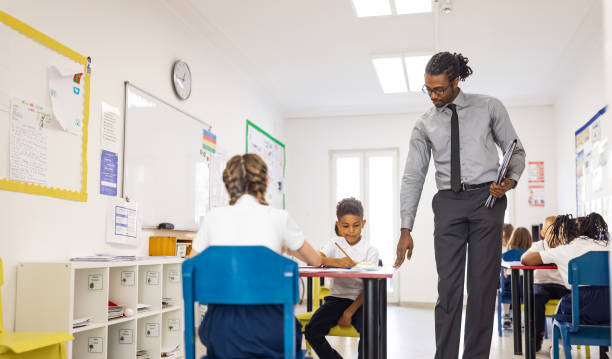 istock Primary School Teacher and students In Classroom. Teacher distributing a educational exam 1318793687