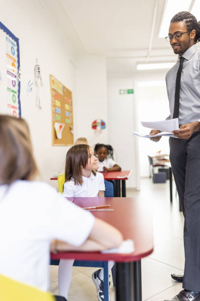 istock Primary School Teacher and students In Classroom. Teacher distributing a educational exam 1318793700