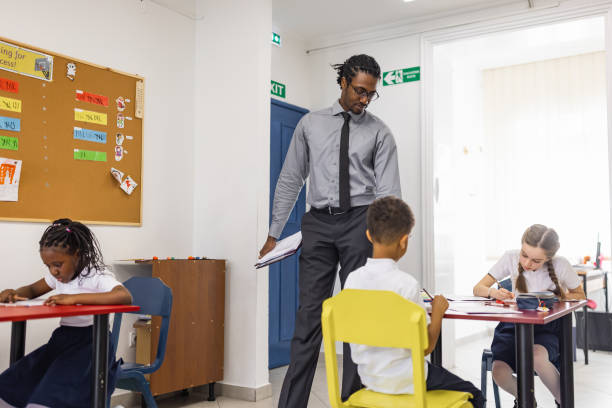 istock Primary School Teacher and students In Classroom. Teacher distributing a educational exam 1318793705