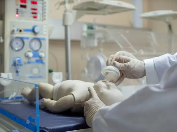 A doctor putting an oxygen mask on a newborn infant model under a radiant warmer/resuscitator unit.