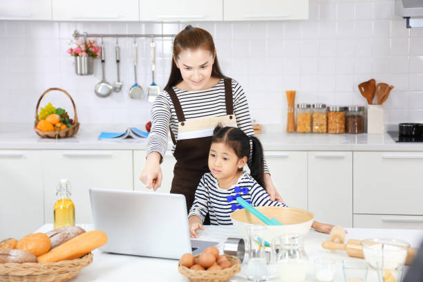 mãe asiática feliz ensinando sua filha a assar pão na cozinha branca moderna olhando para a receita on-line do computador - bread food baked 7 grain bread - fotografias e filmes do acervo