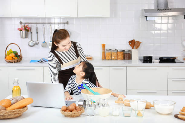feliz madre asiática enseñando a su hija a hornear pan en la cocina moderna blanca mirando la receta en línea desde la computadora - bun bread 7 grain bread dough fotografías e imágenes de stock