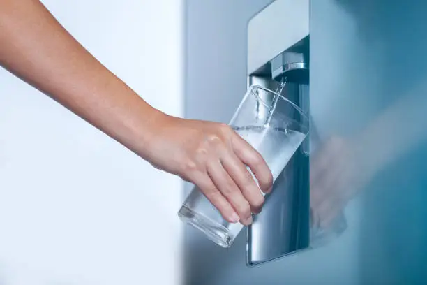 Photo of Water dispenser from dispenser of home fridge, Woman is filling a glass with water from the refrigerator.