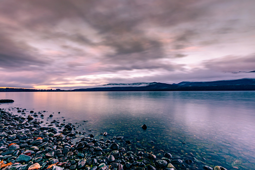 This August 2020 long-exposure image shows the waters of Lake Te Anau in Aotearoa New Zealand at sunrise on a cloudy morning. A few spots of the morning sun shine through the clouds leaving a warm glow. Smooth, multi-coloured stones are seen on the lakeshore.