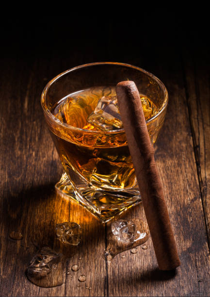 single malt scotch whiskey in modern crystal glass with ice cubes and cuban cigar on wooden background - cigar whisky bar cognac imagens e fotografias de stock