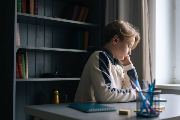 istock Low-angle view of thinking smart schoolboy making notes writing essay in notebook sitting at desk near window 1318820030
