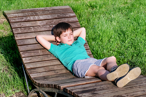 Brunet boy of 8 years in T-shirt and shorts lies on wooden deck chair in the sun against the backdrop of a green lawn. The boy in relaxed, lazy position, closed his eyes.
