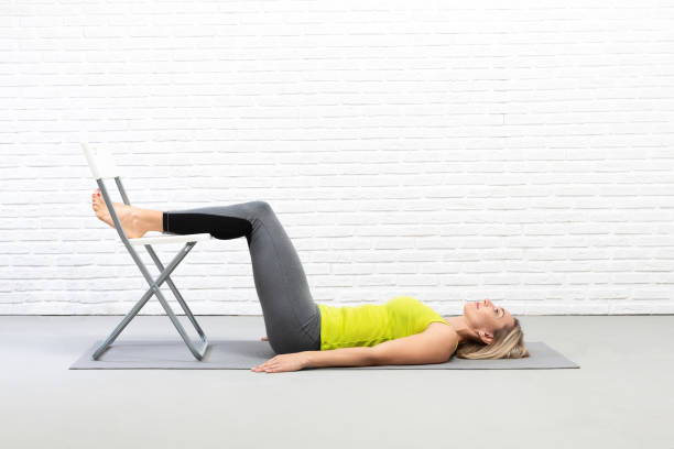 yoga com adereços dentro de casa. mulher adulta caucasiana deita no tapete e coloca os pés na cadeira, relaxa broca, foco seletivo. - female the human body relaxation full length - fotografias e filmes do acervo