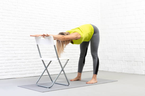 istock Back and hamstrings stretching, yoga indoor with a props. Caucasian woman does deep bend holding on to the chair with her hands. 1318831934