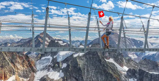 Woman with Swiss flag on Titlis cliff walk suspension bridge. Top of Titlis mountain with glacier in the Uri Alps. Viewpoint 3028 m in cantons of Obwalden and Bern, Switzerland, Europe. Summer season.