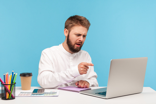 Serious bearded man boss scolding his employees pointing finger at laptop display, conducting online video conference sitting in his office. Indoor studio shot isolated on blue background