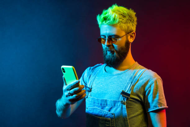 istock Bearded concentrated hipster man holding cell phone in hands, checking social networks, writing new post, has serious expression, wearing denim overalls, 1318839178