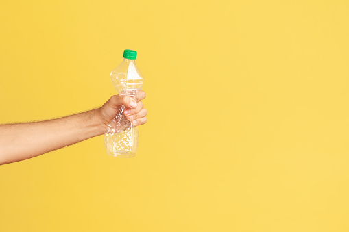 Profile portrait male hand holding and pressing plastic bottle with green cap, sorting his rubbish, worrying about environmental pollution. Indoor studio shot isolated on yellow background