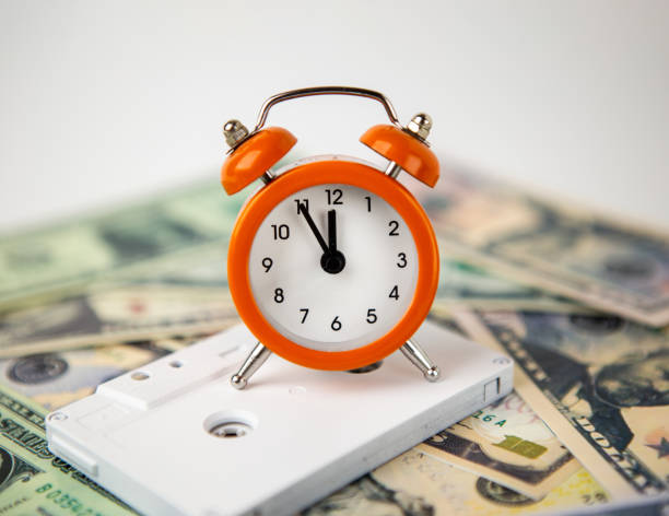 オレンジ色の目覚まし時計は、白いオーディオ音楽カセットと米ドル紙幣に位置しています。時計は5分から12時です。 - time is money audio ストックフォトと画像