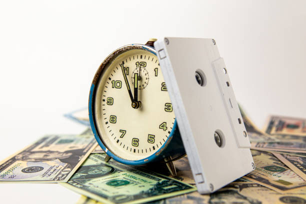 5分から12時を示す古い青い台無しのアナログ時計。時計は米ドル紙幣にあり、その隣には白いオーディオ音楽カセットがあります。時間、お金、音楽。 - time is money audio ストックフォトと画像