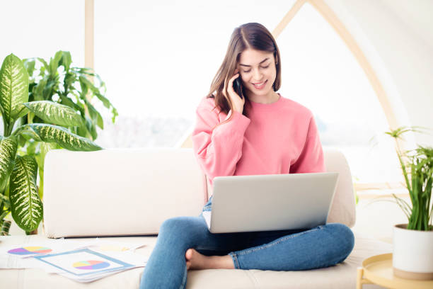 istock Young woman using her mobile phone and laptop while sitting on a couch at home 1318844479
