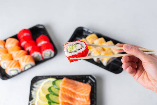 istock Man hands holding california roll with chopsticks. Lunch boxes, sushi set. Assorted rolls philadelphia, sashimi on light background. Restaurant delivery, takeaway concept. Japanese cuisine seafood 1318847484