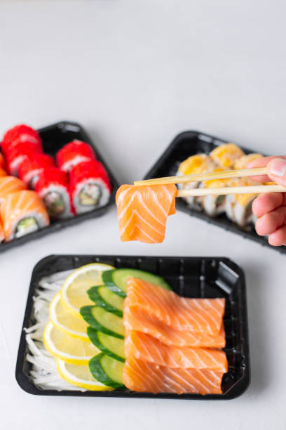 istock Man hands holding raw salmon sashimi chopsticks. Lunch boxes, sushi set. Assorted rolls philadelphia, california, light background. Restaurant delivery, takeaway concept. Japanese cuisine seafood 1318847499
