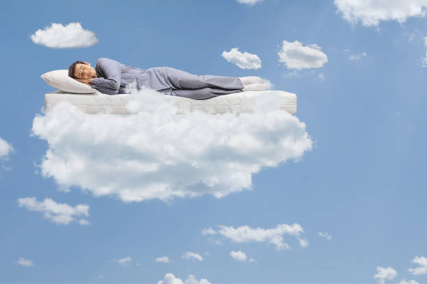 istock Man in pajamas sleeping on a mattress and floating on clouds 1318858138