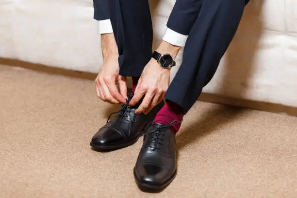 A businessman in dark blue suit, red socks and watch tying his shoelaces of black shoes. Modern, stylish and expensive look for young man