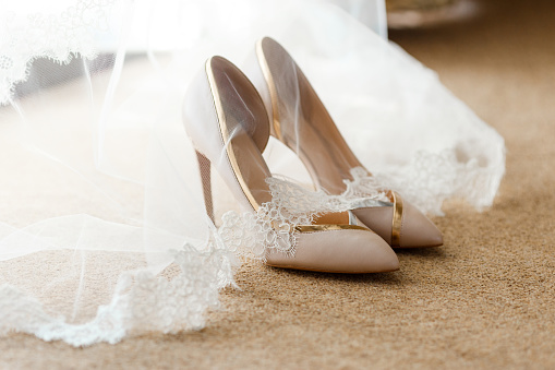 Beige bridal shoes standing on the floor, covered with a white veil with lace. A pair of expensive and beautiful wedding shoes with high heels, with gold and silver finish