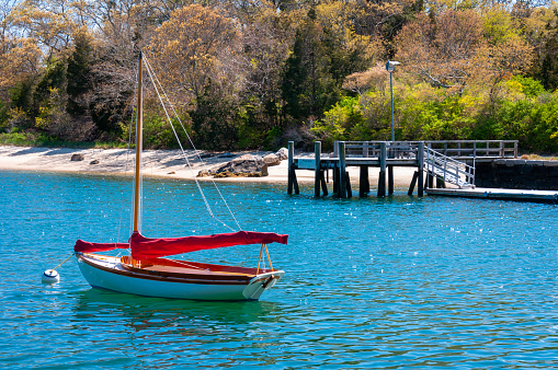 A small catboat with a red sail covering rests, moored in Quissett Harbor, in Falmouth, Massachusetts.