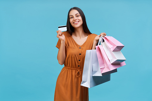 Studio shot of beautiful happy smiling brunette girl posing with pile of shopping bags and credit card in hands, isolated on pastel blue background.