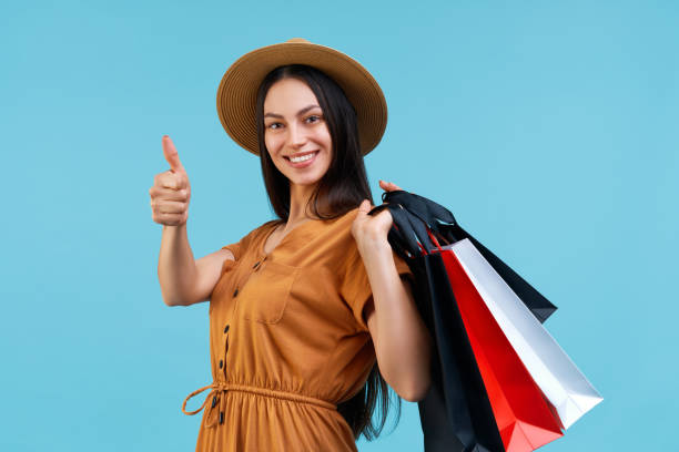 Beautiful smiling confident young woman in trendy boho-style dre Beautiful smiling confident young woman in trendy boho-style dress and boater hat, holding shopping bags in hand and showing thumbs up gesture, isolated over bright blue background. dre stock pictures, royalty-free photos & images