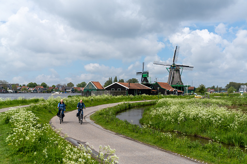 Zaandam, Netherlands - 18 May, 2021: Dutch senior citizens enjoy a bicycle ride along the canals of North Holland with traditional windmills in the background