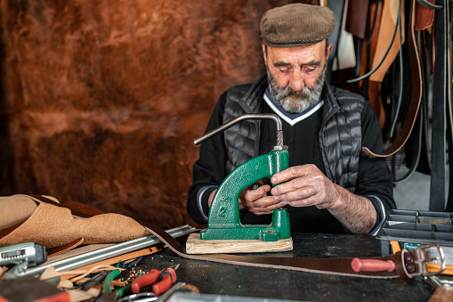Senior craftsman making holes in leather with puncher. Hairy leather background.