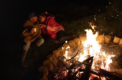 Rural Latin American couple on a romantic date having at a bonfire and eating marshmallows while drinking coffee and kissing