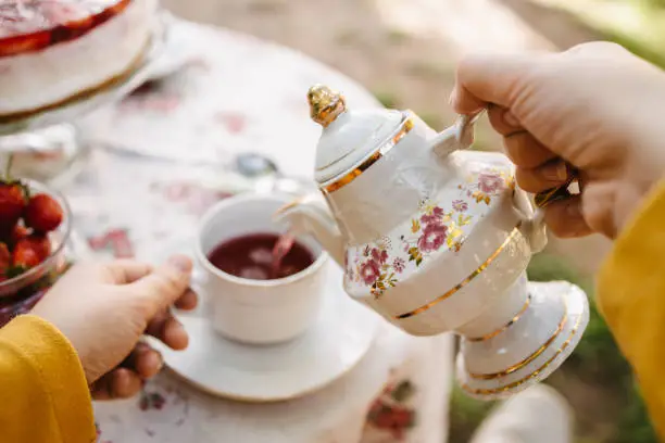 A close-up of a young adult woman pouring tea out of a teapot into a teacup during the tea party in the garden.