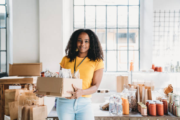 portrait of a mixed race volunteer woman holding a cardboard box of food and drink at the food bank - food donation box groceries canned food imagens e fotografias de stock