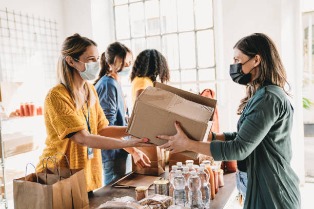 A millennial woman is taking a box of food and drink at the food and clothes bank A millennial woman is taking a box of food and drink at the food and clothes bank. People are wearing protective face masks. charitable foundation stock pictures, royalty-free photos & images