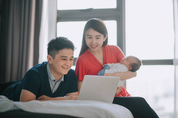 asiatische chinesische vater liegt auf dem bett mit laptop online-shopping, während mutter trägt neugeborenen baby junge im schlafzimmer - women group of people lying down mother stock-fotos und bilder