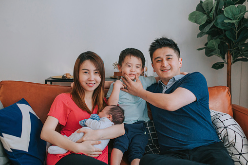 portrait asian chinese young family with 2 children looking at camera smiling sitting on sofa in living room