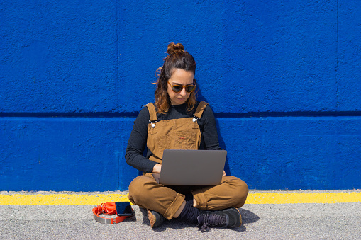 Lifestyle woman with sunglasses sitting on the ground and leaning against the wall on the street on a sunny day taking a selfie and working with laptop and headphones by her side.