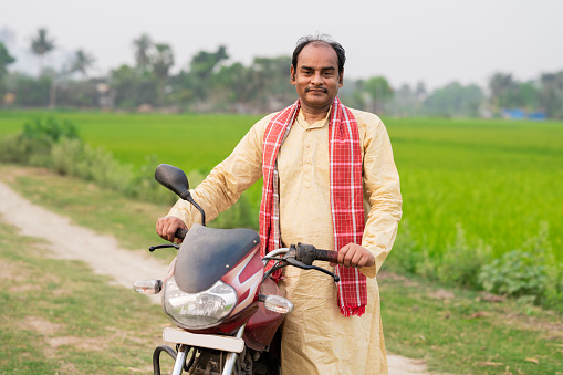 Outdoor image of Middle aged Indian farmer standing with his motorbike in green agricultural field.