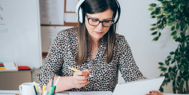 istock Female Teacher or Student with Headset Holding Tablet and Writing Notes in Papers 1318895444