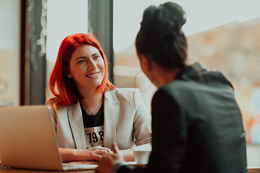 One-on-one meeting.Two young business women sitting at table in cafe. Girl shows colleague information on laptop screen. Girl using smartphone, blogging. Teamwork, business meeting. Freelancers working.