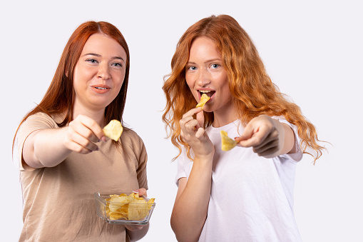 Close up of two ginger, red-haired female girls, friends holding bowl with fresh crispy potato chips sharing with friends at home party. Showing snack chips to camera, smiling. High quality photo