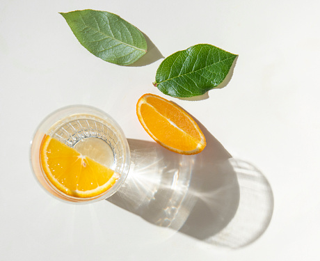 A glass of mineral water and a slice of orange on a light  table with fresh juicy fruit oranges and green leaves with hard shadows. Summer time. Top view image
