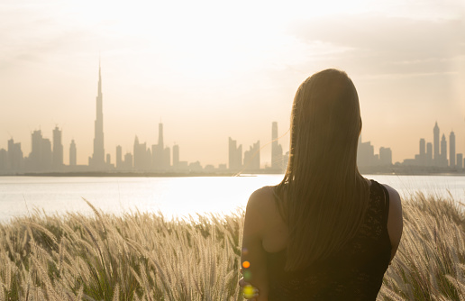 Young woman standing looking at the beautiful Dubai cityscape skyline view on the waterfront during sunset.
