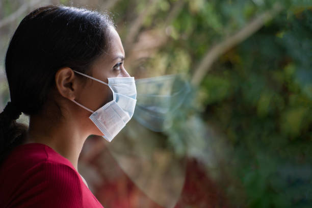 joven mujer triste con una máscara facial mirando a través de la ventana con su reflejo en el vidrio, auto aislado en casa. - andres fotografías e imágenes de stock