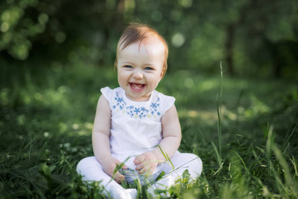 das baby sitzt auf dem grünen gras im park und lacht. die ersten beiden zähne sind sichtbar, wenn das baby lächelt. gesundes baby lacht. pflege, pflege, - cute girl stock-fotos und bilder