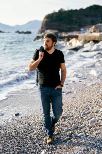 istock Pensive man walking along a pebble beach with a denim jacket over his shoulder and a hand in his pocket against the background of the sea and rocks 1318929937